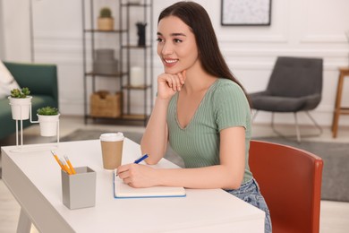 Young woman writing in notebook at white table indoors