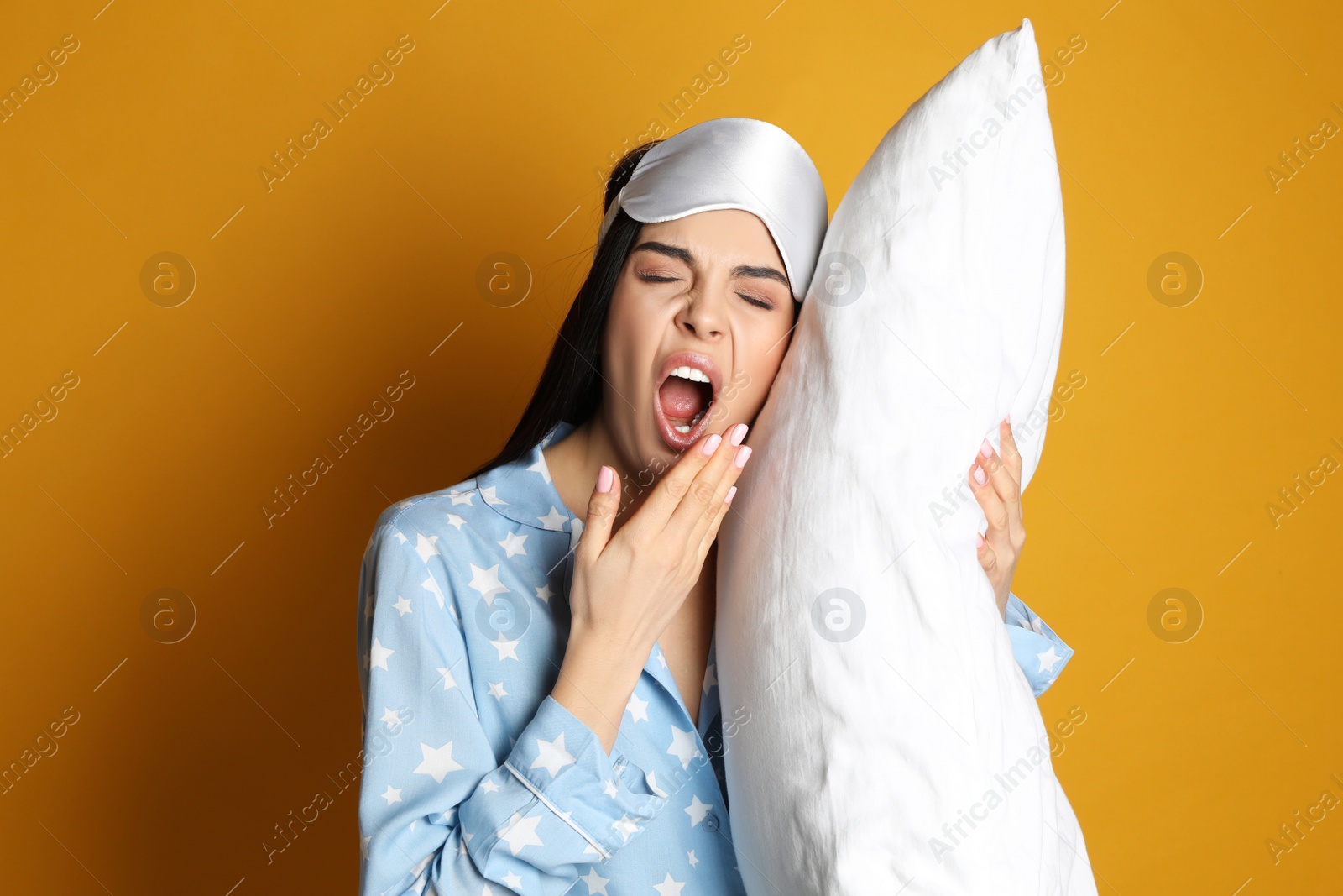 Photo of Young tired woman with sleeping mask and pillow yawning on yellow background