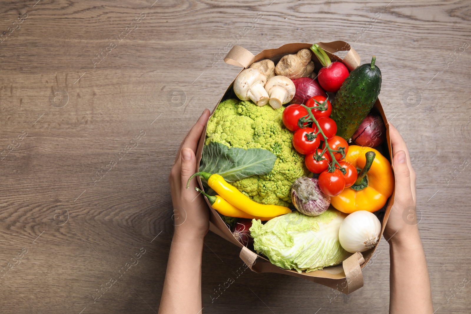 Photo of Woman holding paper package full of fresh vegetables on wooden background, top view. Space for text