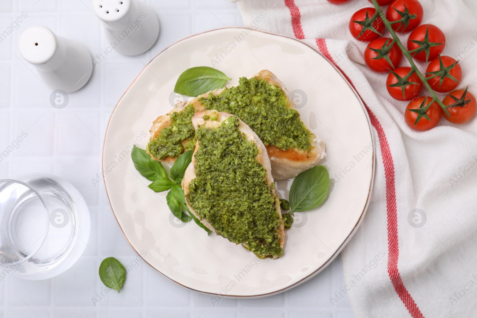 Photo of Delicious chicken breasts with pesto sauce, tomatoes and glass of water on white tiled table, flat lay