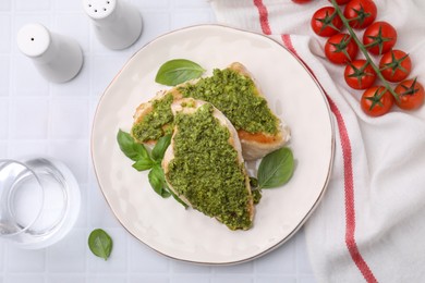 Delicious chicken breasts with pesto sauce, tomatoes and glass of water on white tiled table, flat lay