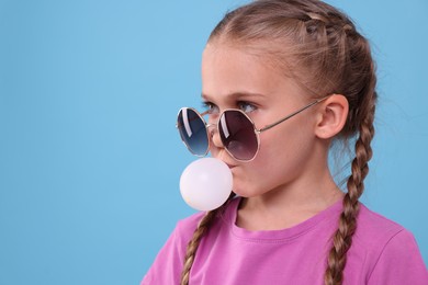 Photo of Girl in sunglasses blowing bubble gum on light blue background, space for text