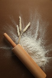 Flour, spikelets and rolling pin on brown table, top view