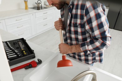 Photo of Young man using plunger to unclog sink drain in kitchen, closeup
