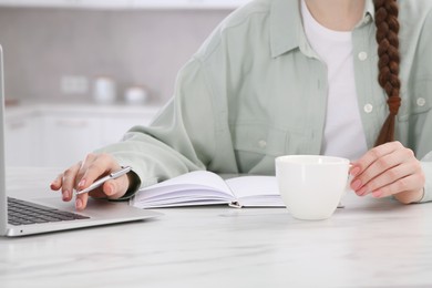 Woman with cup of coffee and notebook working on laptop at white marble table in kitchen, closeup