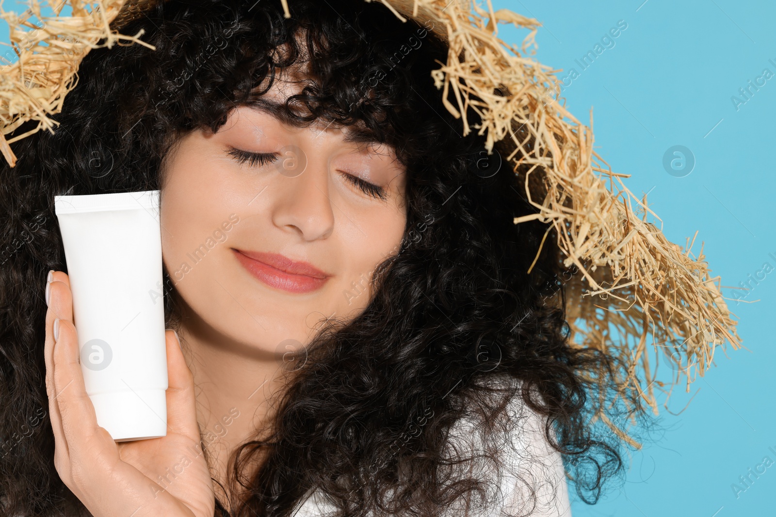 Photo of Beautiful young woman in straw hat holding tube of sun protection cream on light blue background, closeup