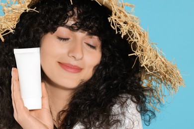 Beautiful young woman in straw hat holding tube of sun protection cream on light blue background, closeup