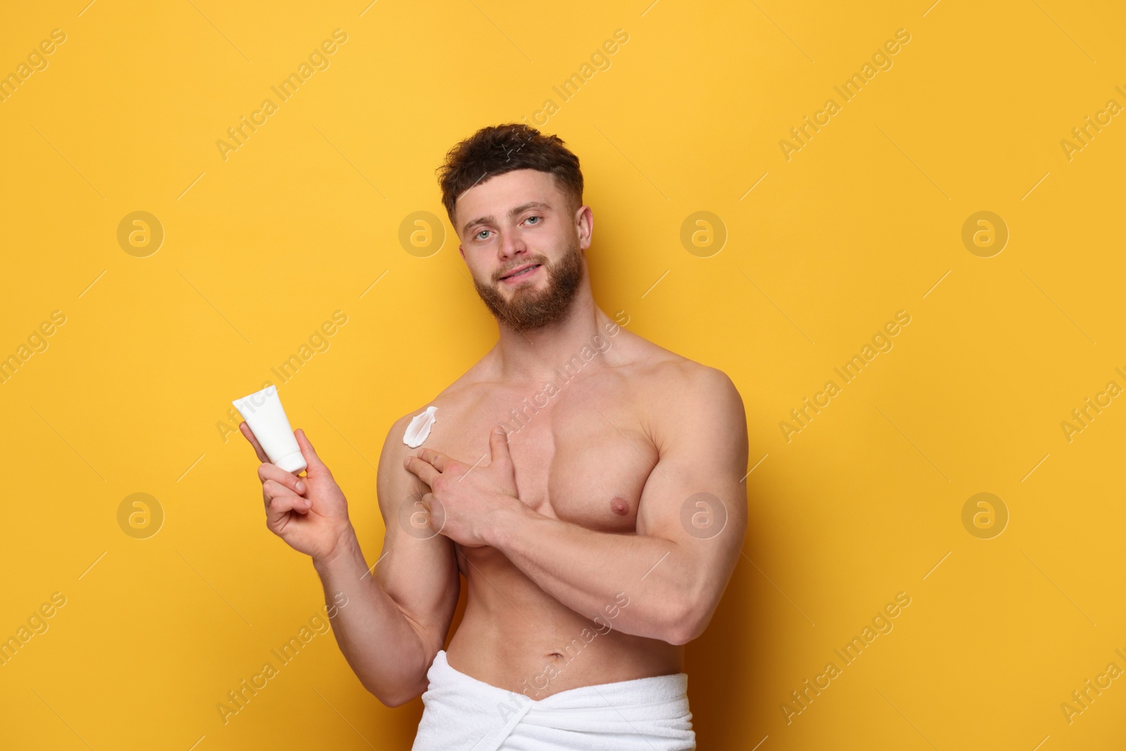 Photo of Handsome man applying moisturizing cream onto his shoulder on orange background