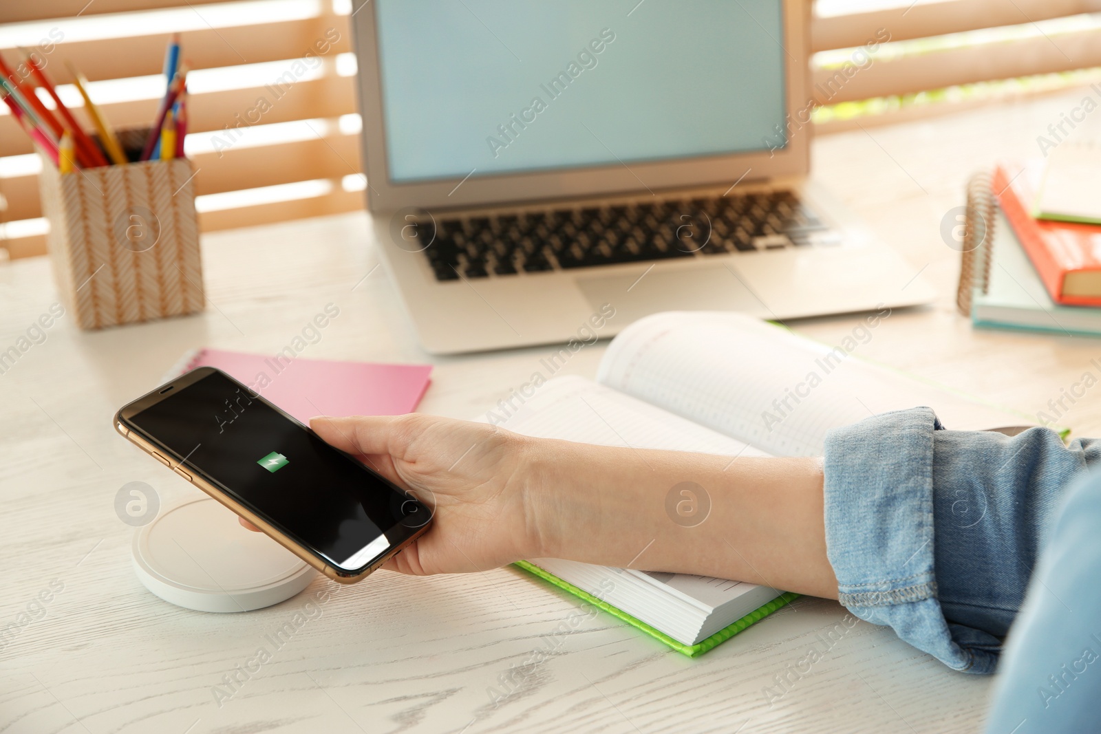 Photo of Woman putting mobile phone onto wireless charger at white wooden table, closeup. Modern workplace accessory