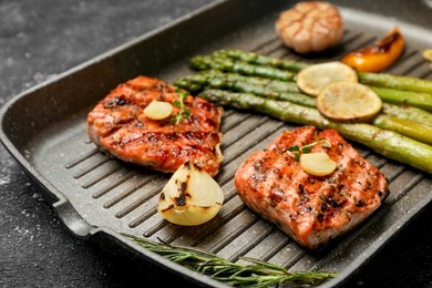Frying pan with tasty grilled salmon, lemon and asparagus on black table, closeup