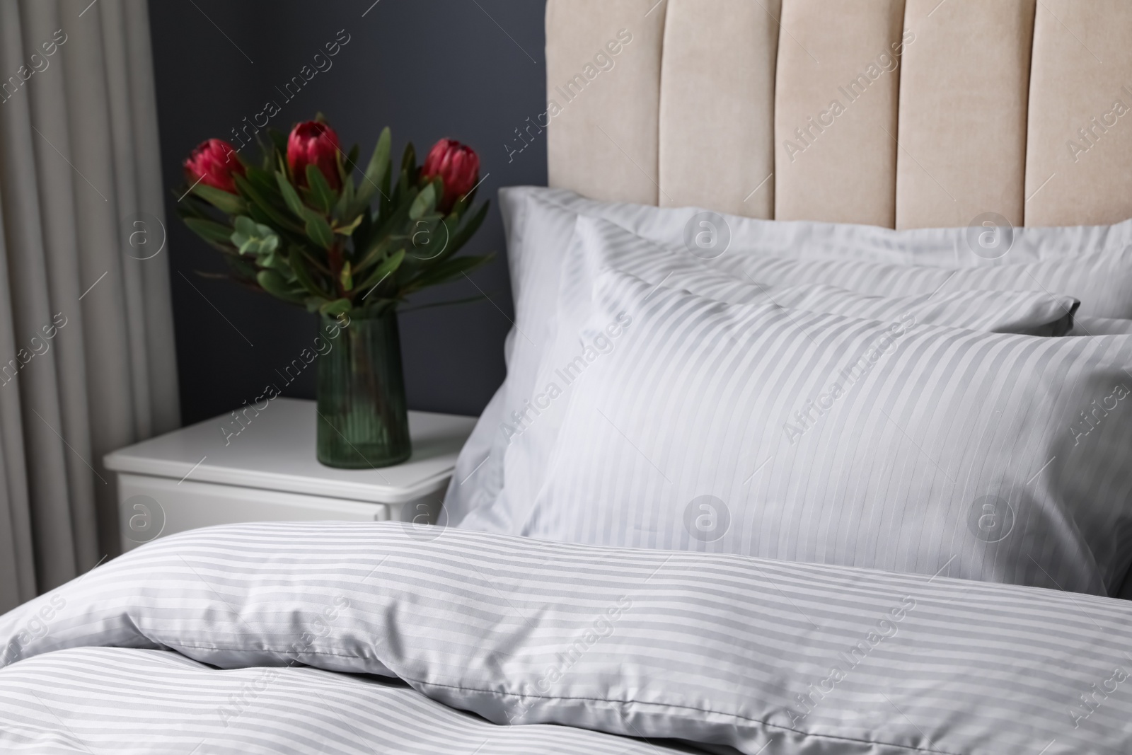 Photo of Nightstand and flowers near comfortable bed with soft pillows indoors