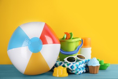Colorful beach ball, other accessories and sand toys on light blue wooden table against yellow background