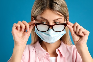 Woman with foggy glasses caused by wearing disposable mask on blue background. Protective measure during coronavirus pandemic