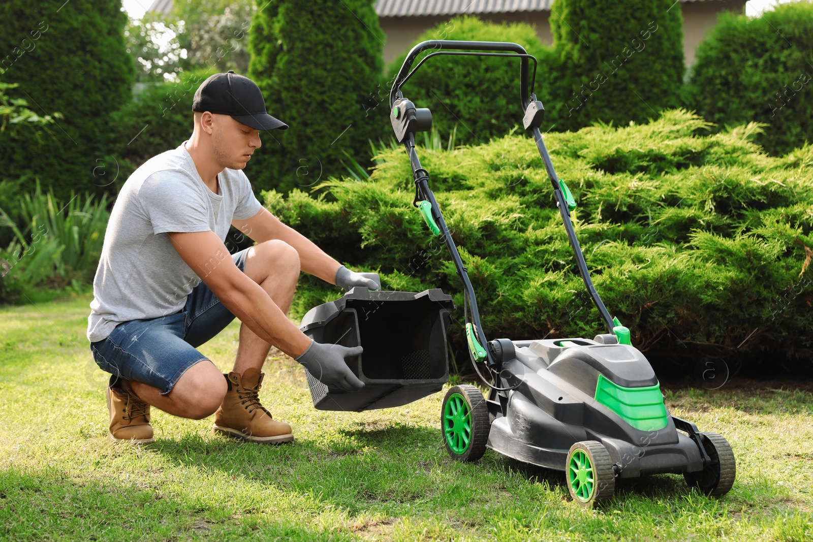 Photo of Cleaning lawn mower. Young man detaching grass catcher from device in garden