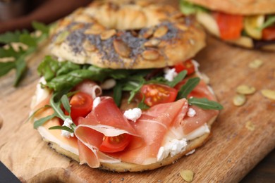 Photo of Tasty bagel with cured ham, cream cheese, tomatoes and arugula on wooden board, closeup