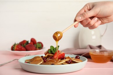 Photo of Woman pouring honey onto cereal pancakes with berries on pink wooden table, closeup