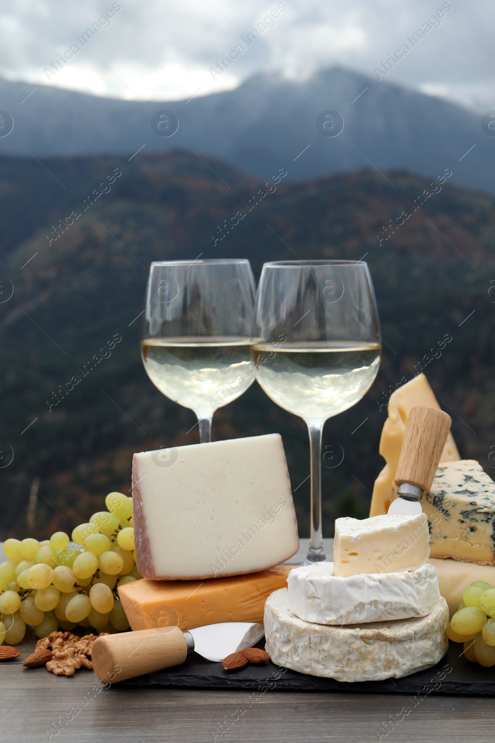 Photo of Different types of delicious cheeses, snacks and wine on wooden table against mountain landscape