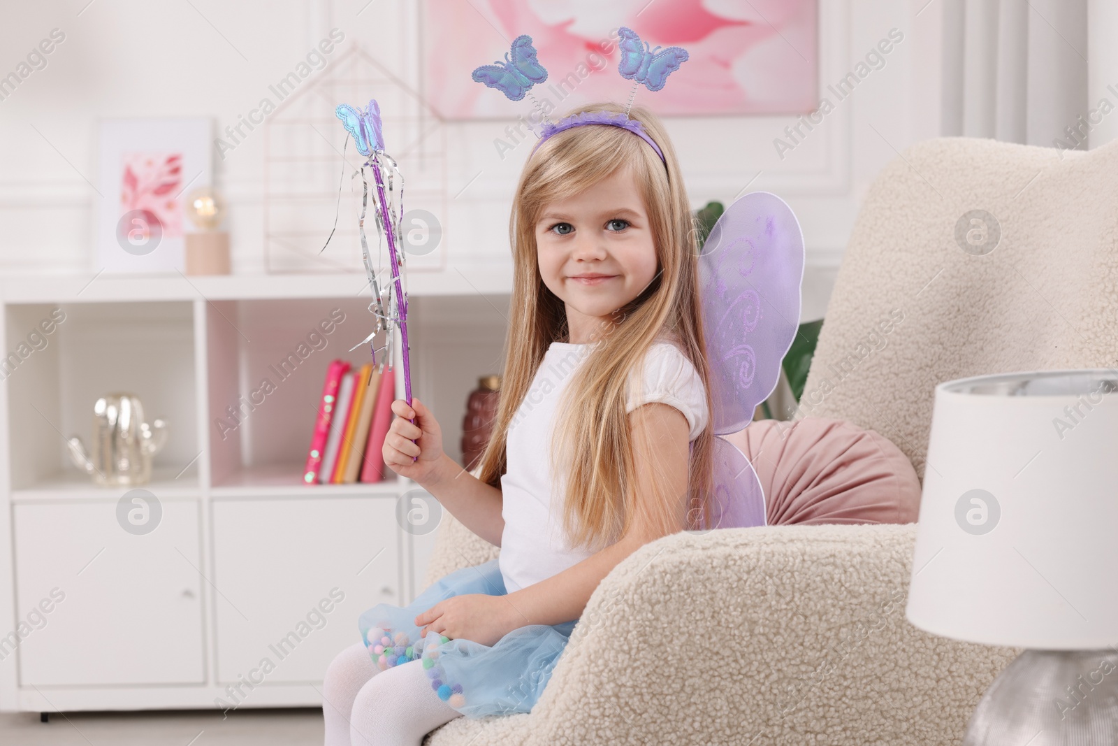 Photo of Cute little girl in fairy costume with violet wings and magic wand in armchair at home, space for text