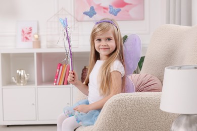 Cute little girl in fairy costume with violet wings and magic wand in armchair at home, space for text