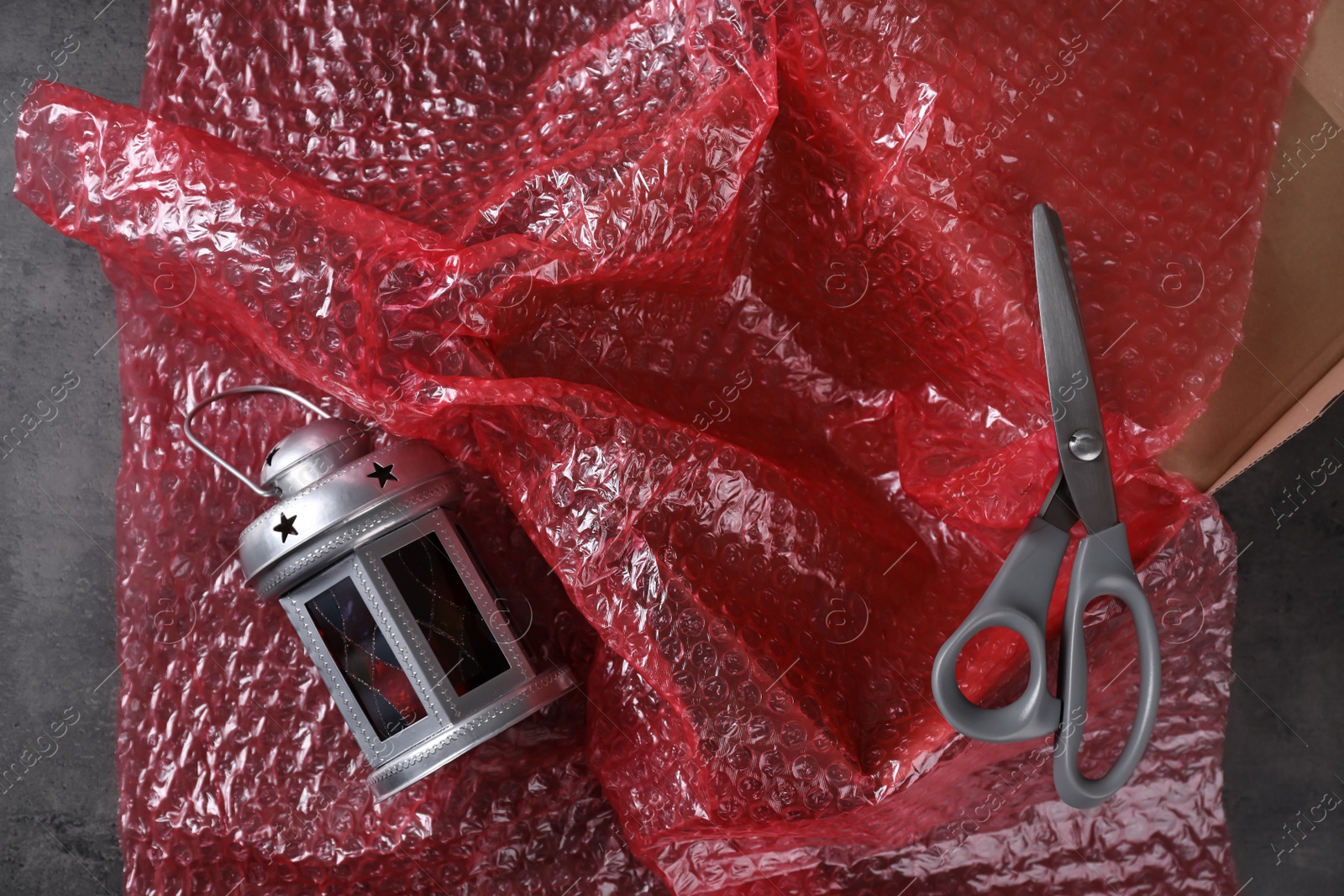 Photo of Beautiful decorative lantern, red bubble wrap, scissors and cardboard box on dark grey table, flat lay