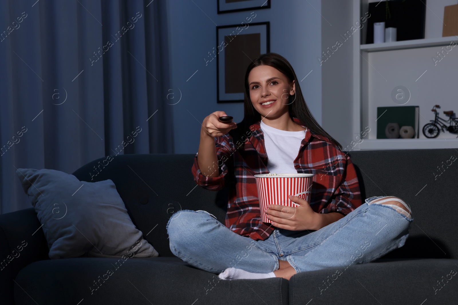 Photo of Happy woman holding popcorn bucket and changing TV channels with remote control at home in evening