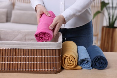 Photo of Woman putting rolled shirt into basket at table in room, closeup. Organizing clothes