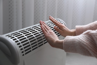 Photo of Woman warming hands near electric heater at home, closeup