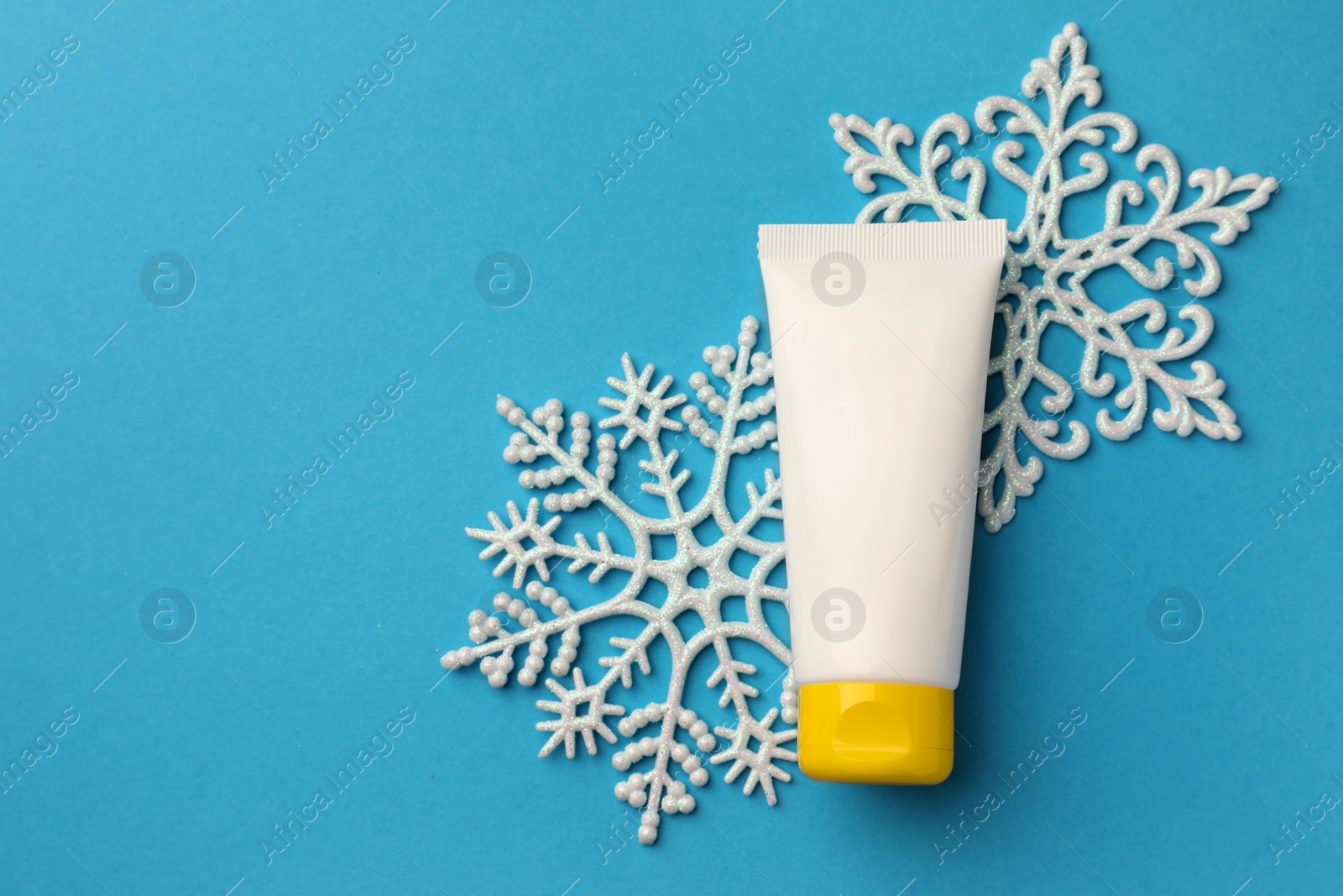 Photo of Tube of hand cream and snowflakes on light blue background, flat lay with space for text. Winter skin care