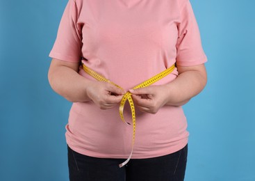 Overweight woman measuring waist with tape on light blue background, closeup