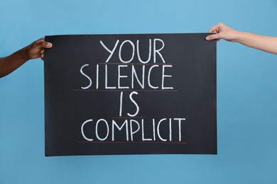 Photo of Woman and African American man holding sign with phrase Your Silence Is Complicit on light blue background, closeup. Racism concept