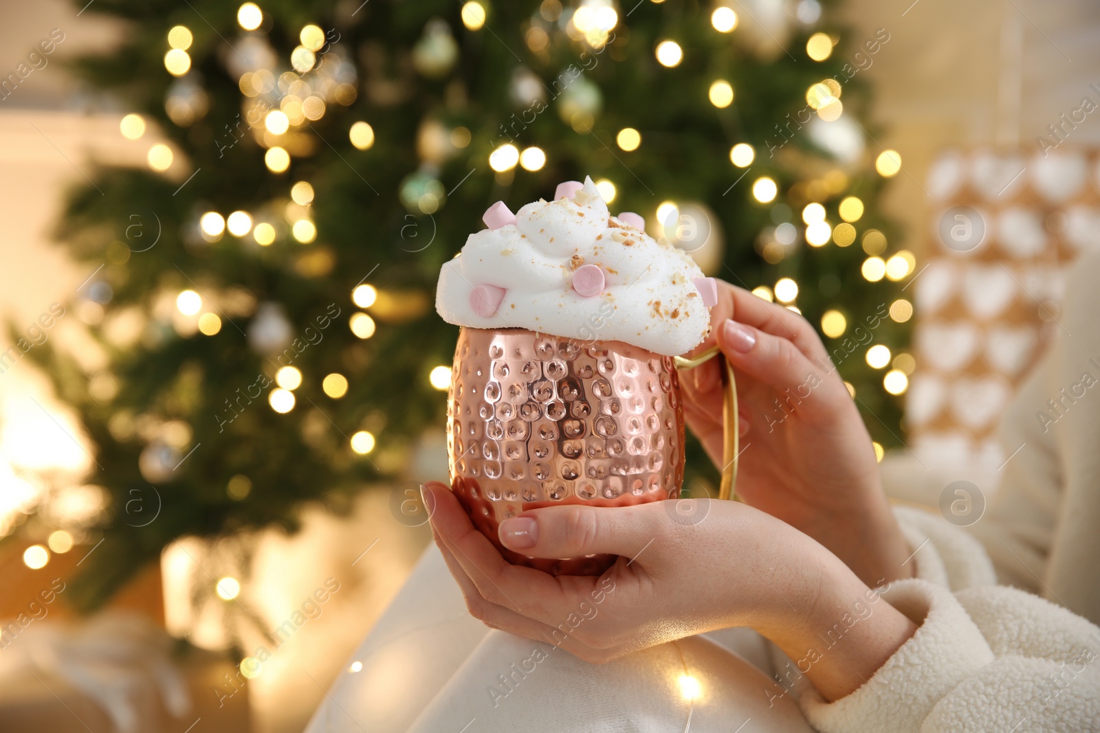 Photo of Woman holding cup of delicious drink with whipped cream and marshmallows near Christmas tree indoors, closeup