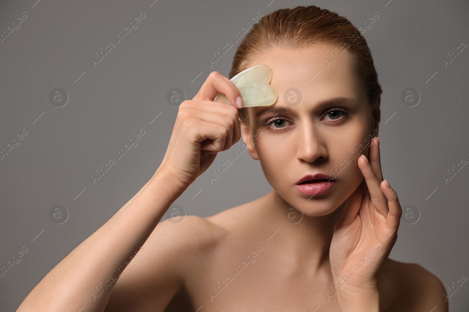 Photo of Beautiful young woman doing facial massage with gua sha tool on grey background