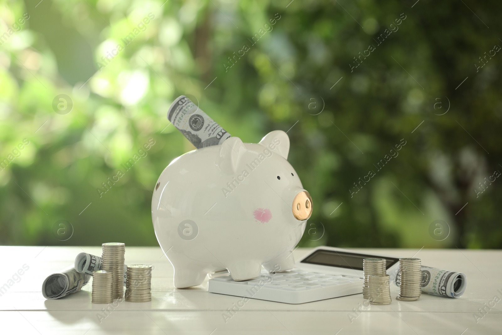 Photo of Financial savings. Piggy bank with dollar banknotes, stacked coins and calculator on white wooden table outdoors