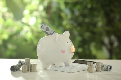 Photo of Financial savings. Piggy bank with dollar banknotes, stacked coins and calculator on white wooden table outdoors