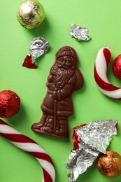 Chocolate Santa Claus among sweets on light green background, flat lay