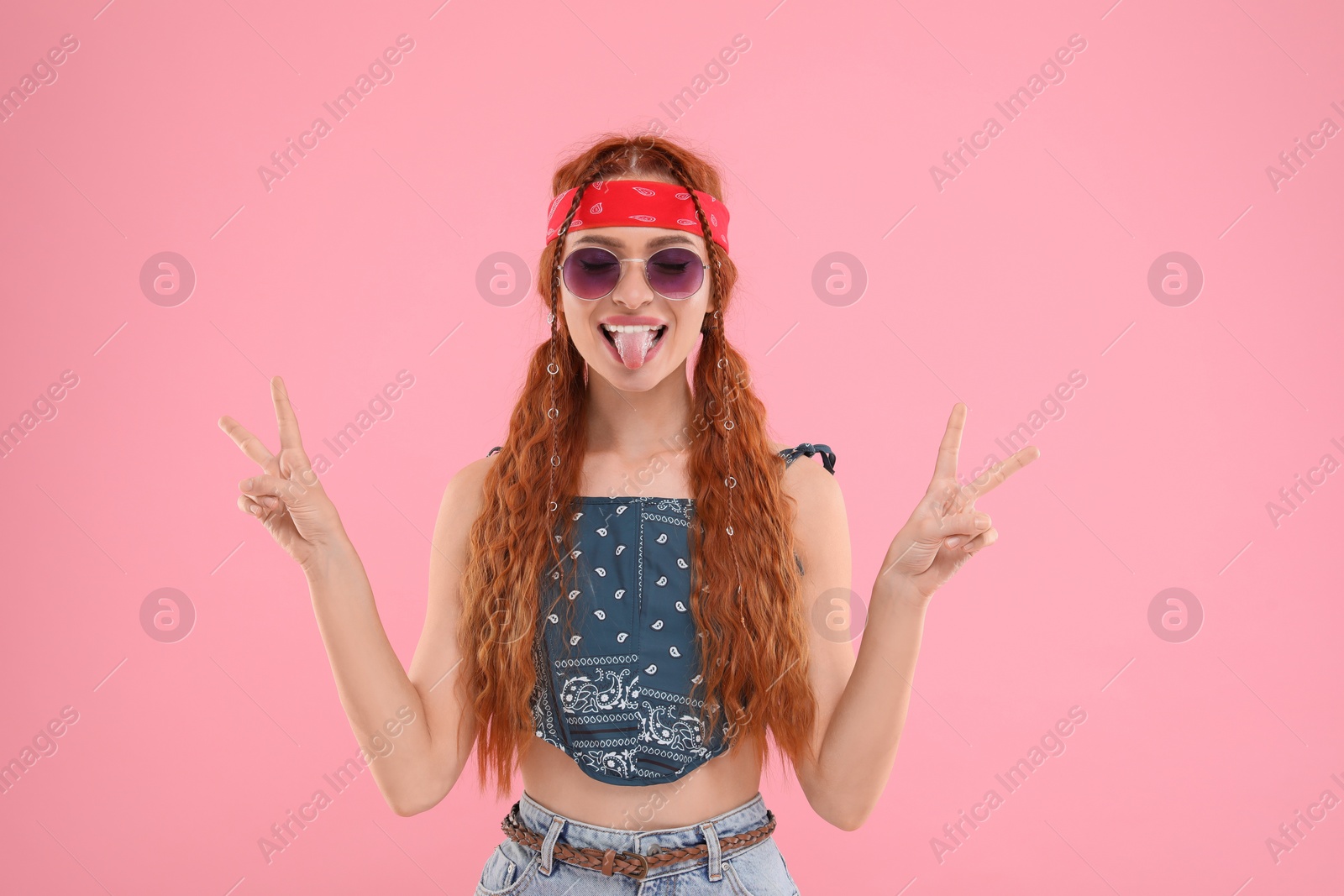 Photo of Stylish young hippie woman in sunglasses showing V-sign on pink background