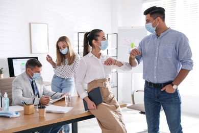 Photo of Coworkers with protective masks making elbow bump in office. Informal greeting during COVID-19 pandemic