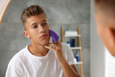 Photo of Young man washing his face with sponge near mirror in bathroom