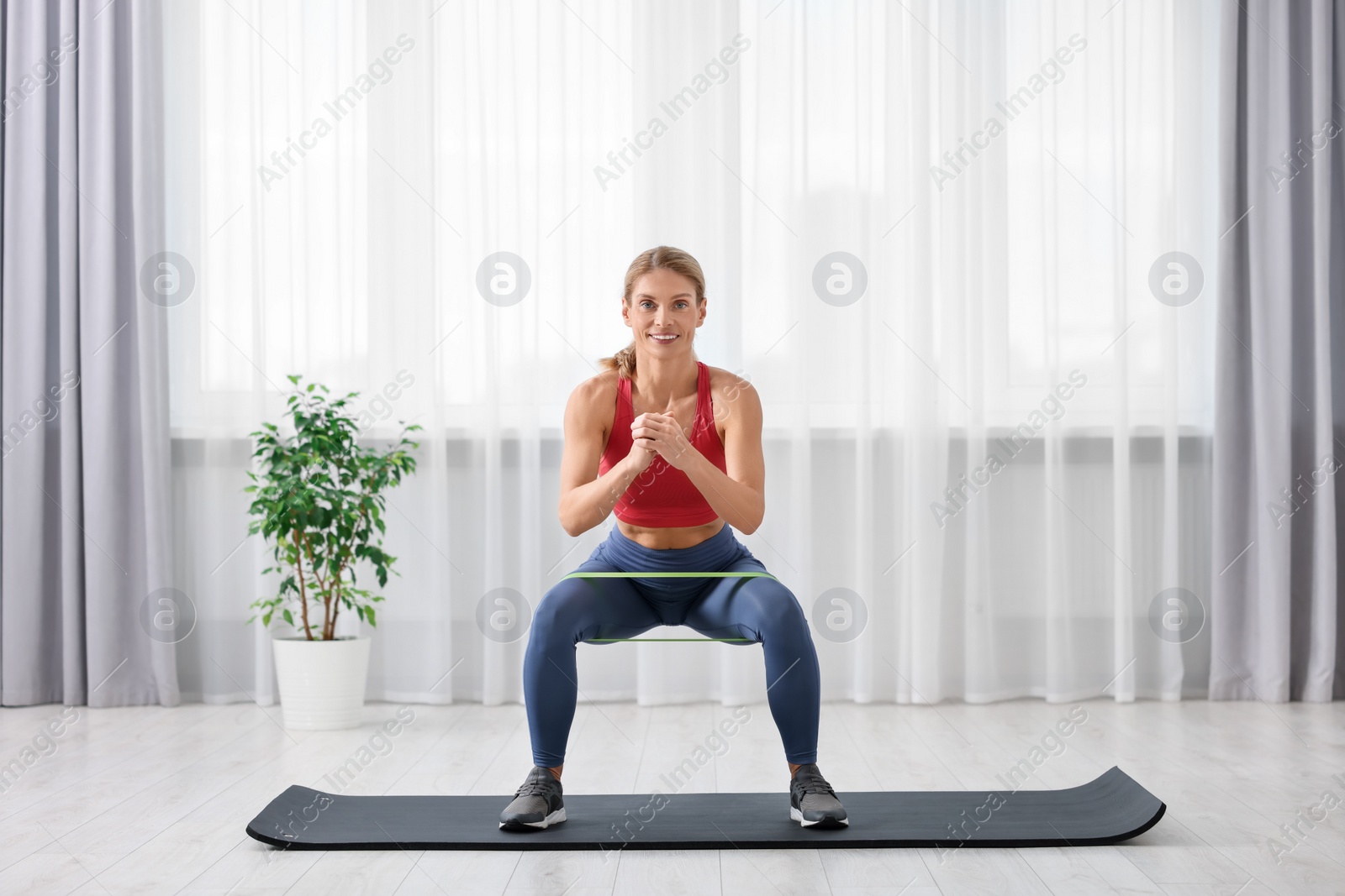 Photo of Athletic woman doing squats with fitness elastic band on mat at home