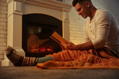 Man warming up with orange plaid while reading book at fireplace