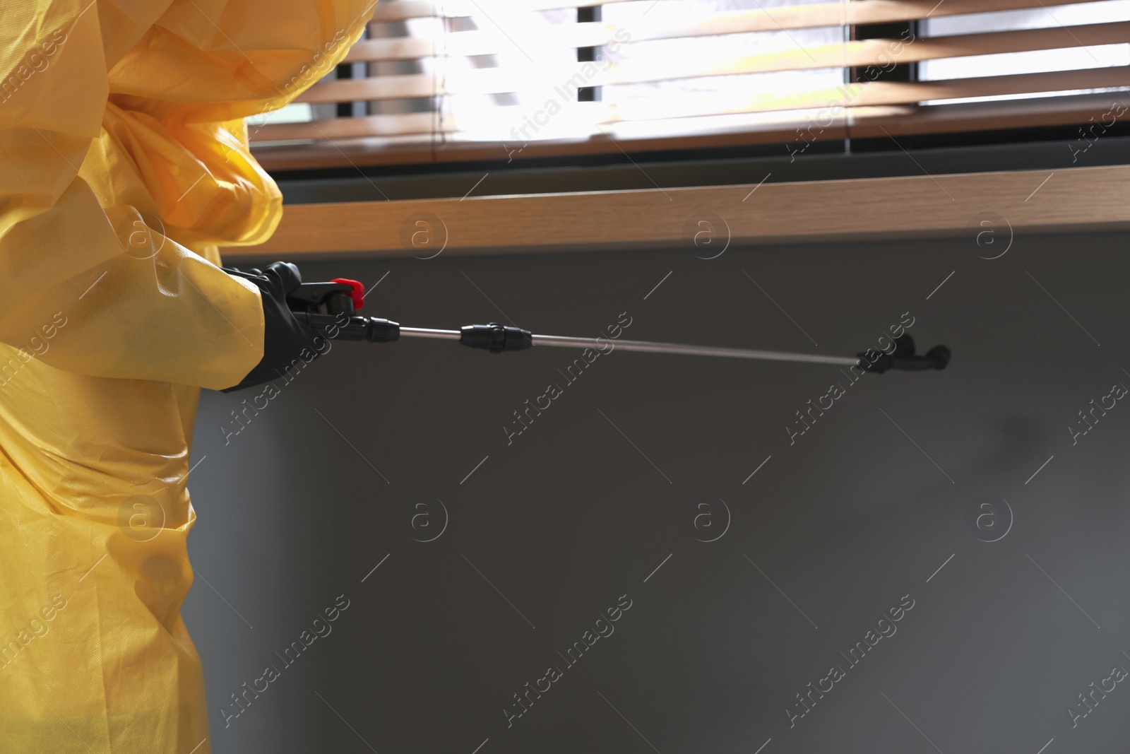 Photo of Pest control worker in protective suit spraying insecticide on window sill indoors, closeup