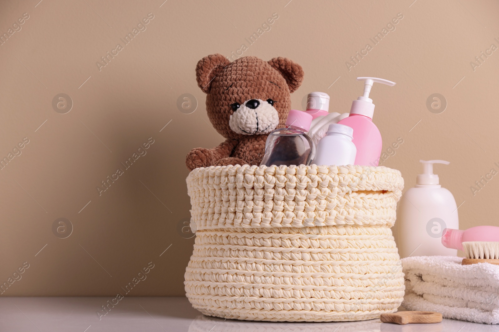 Photo of Knitted basket with baby cosmetic products, bath accessories and toy bear on white table against beige background. Space for text