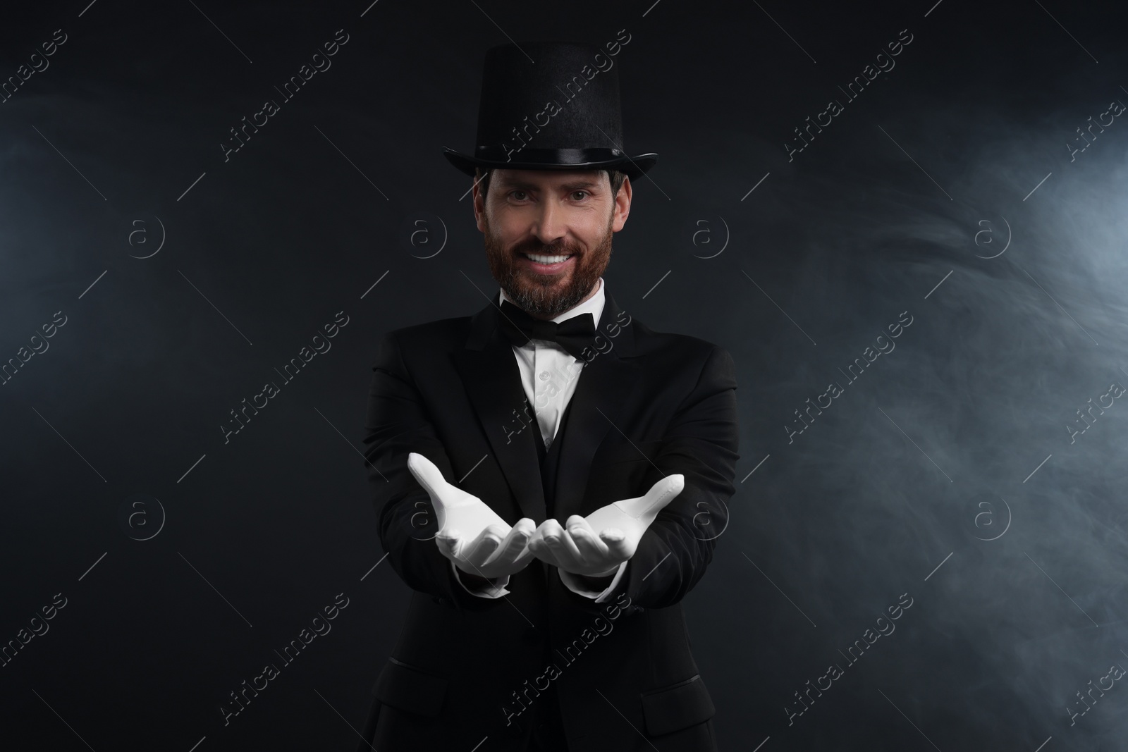 Photo of Happy magician wearing top hat in smoke on black background