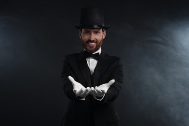 Happy magician wearing top hat in smoke on black background