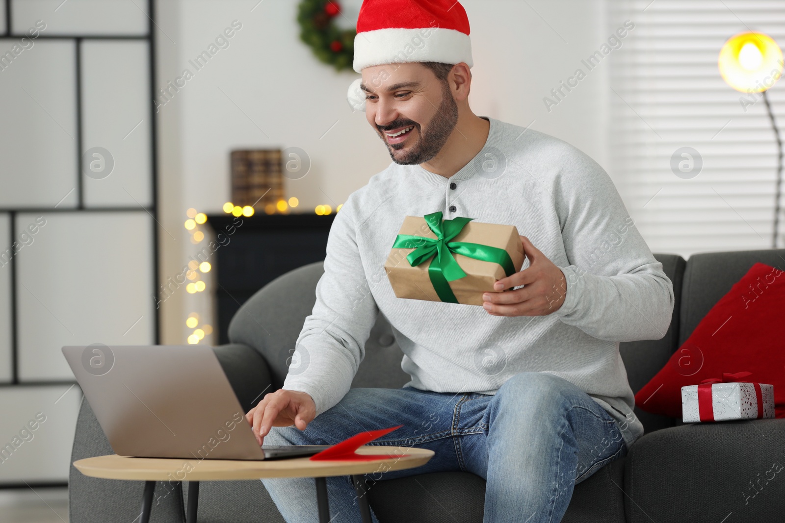 Photo of Celebrating Christmas online with exchanged by mail presents. Happy man in Santa hat with gift box during video call on laptop at home