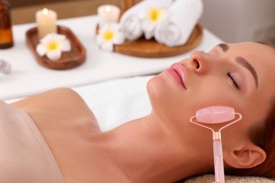 Photo of Young woman receiving facial massage with rose quartz roller in beauty salon, closeup