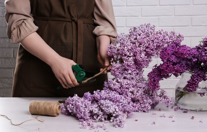 Woman trimming lilac branches with secateurs at white wooden table, closeup
