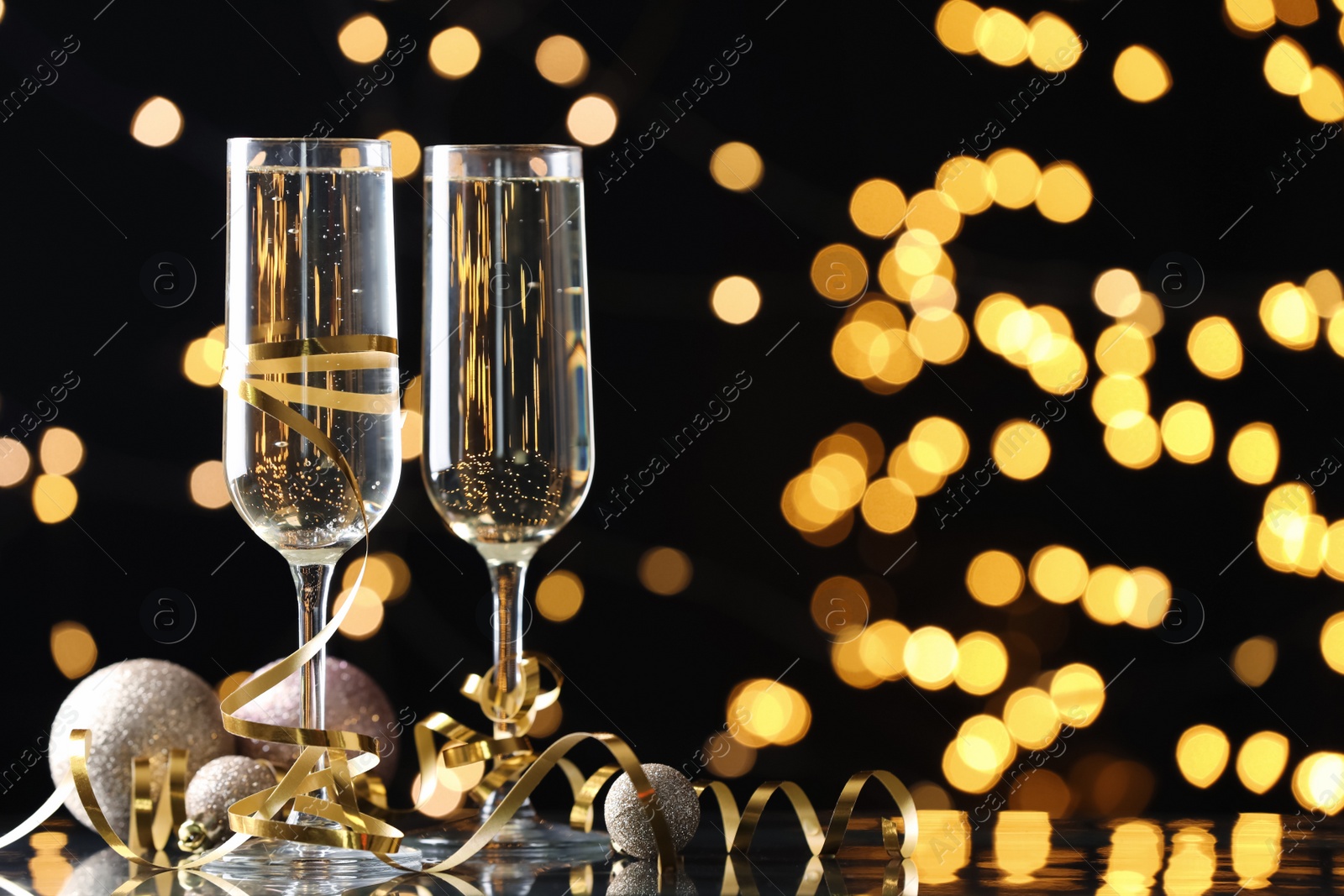 Photo of Glasses of champagne, Christmas decor and serpentine streamers against black background with blurred lights. Space for text