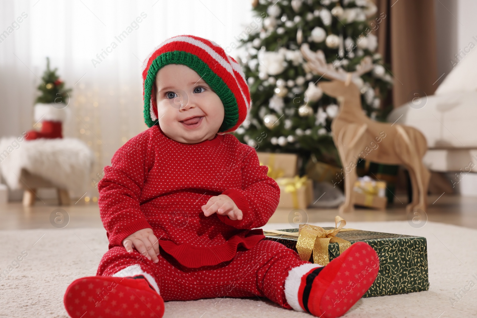 Photo of Baby in elf hat near Christmas gift on floor at home