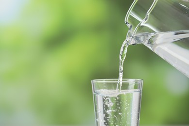 Photo of Pouring fresh water from jug into glass against blurred green background, closeup. Space for text
